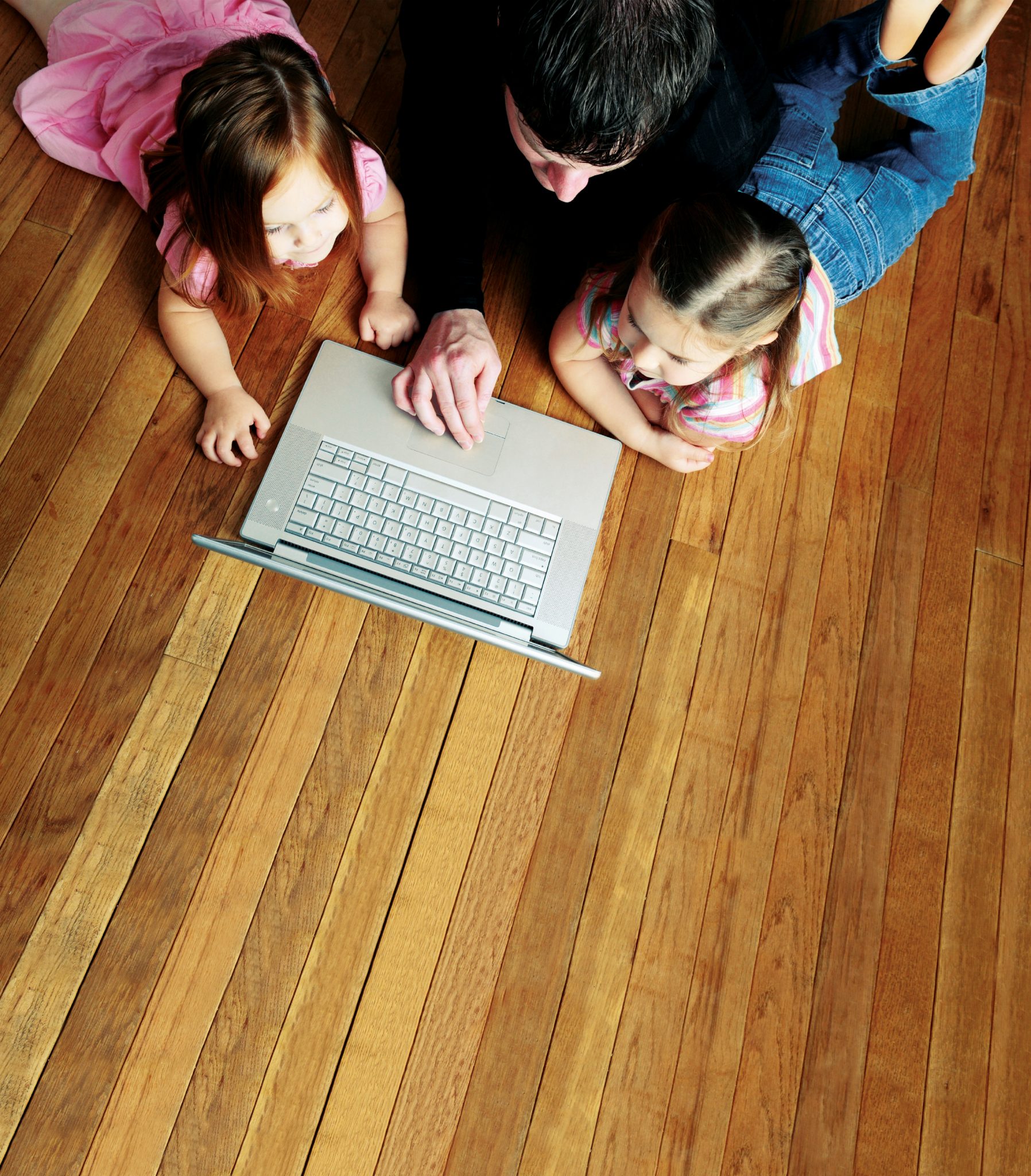 A dad and two girls look lie on a hardwood floor and watch something on a laptop