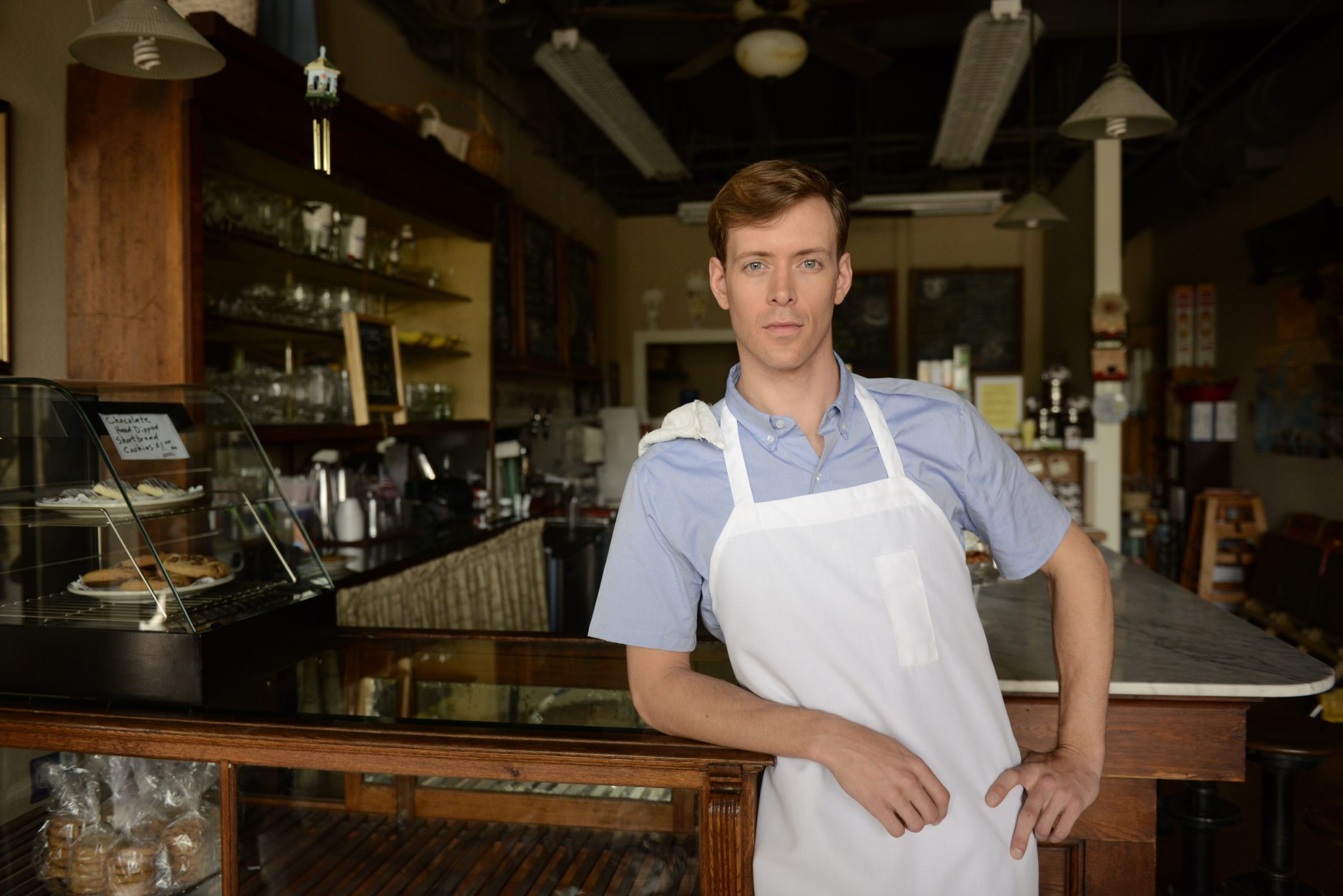 A coffee shop owner wears and apron and leans on the countertop