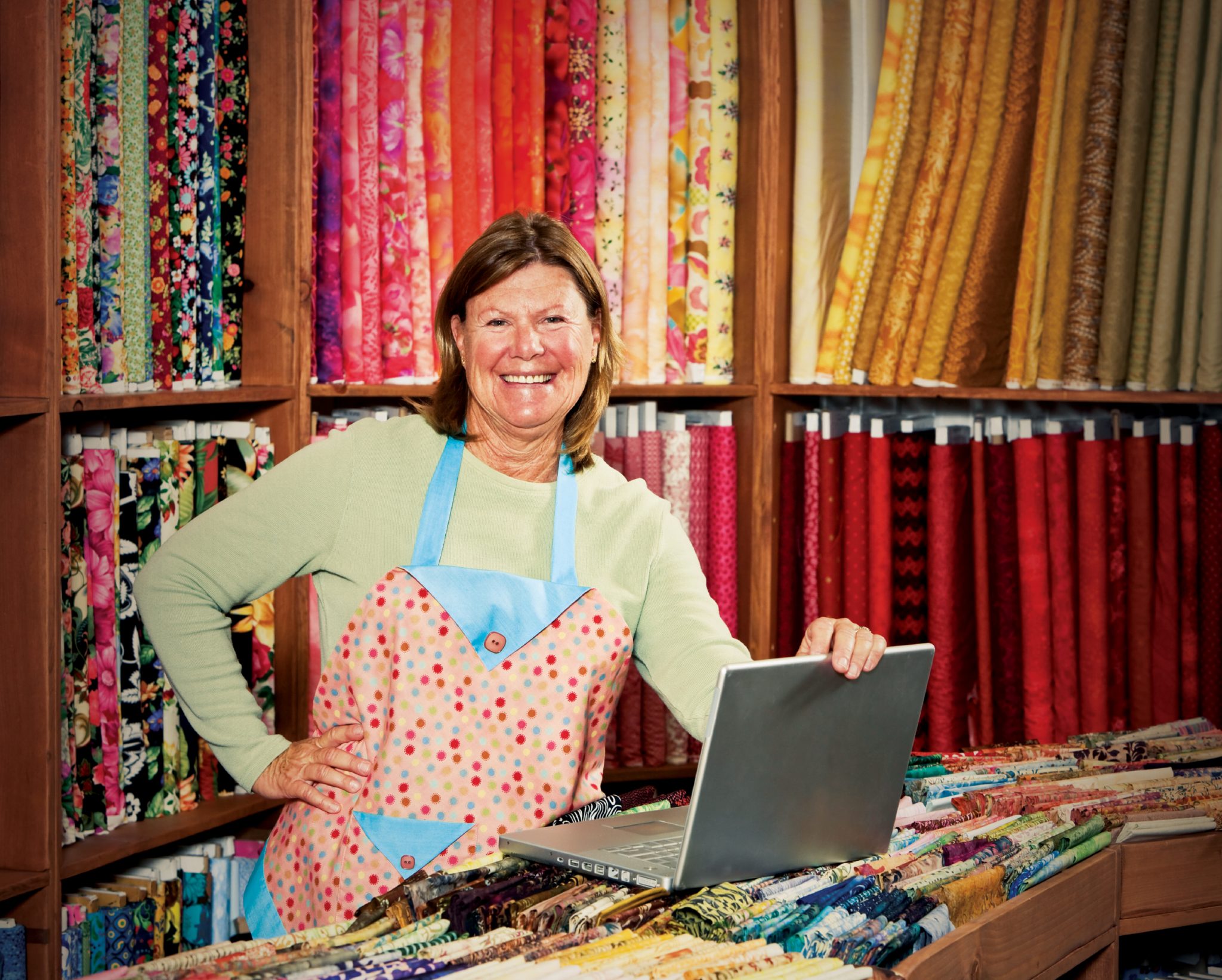 A Quilt Shop Owner uses her laptop at her store surrounded by bolts of fabric