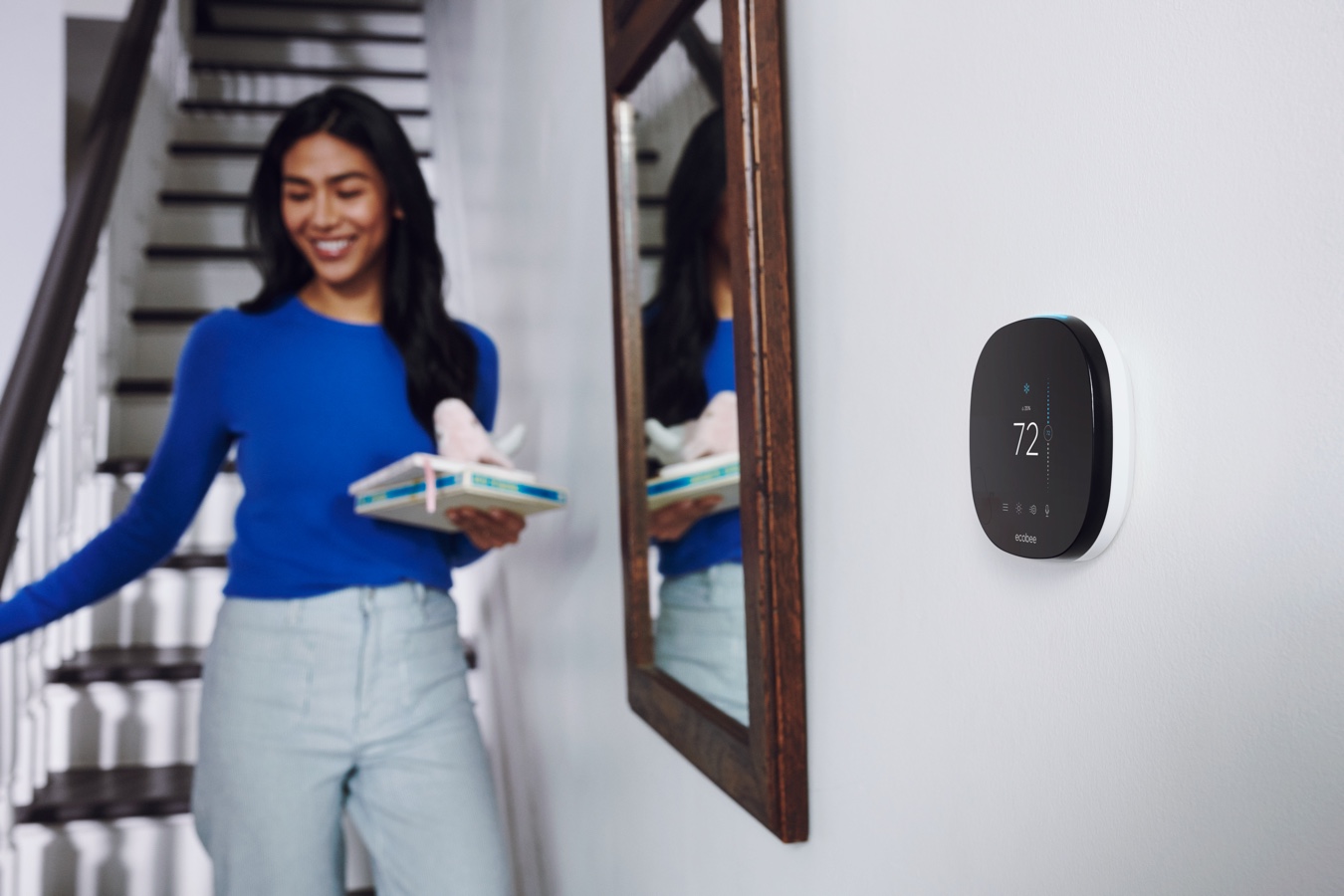 A woman walks down stairs in her home. There is a smart thermostat on the wall near her.