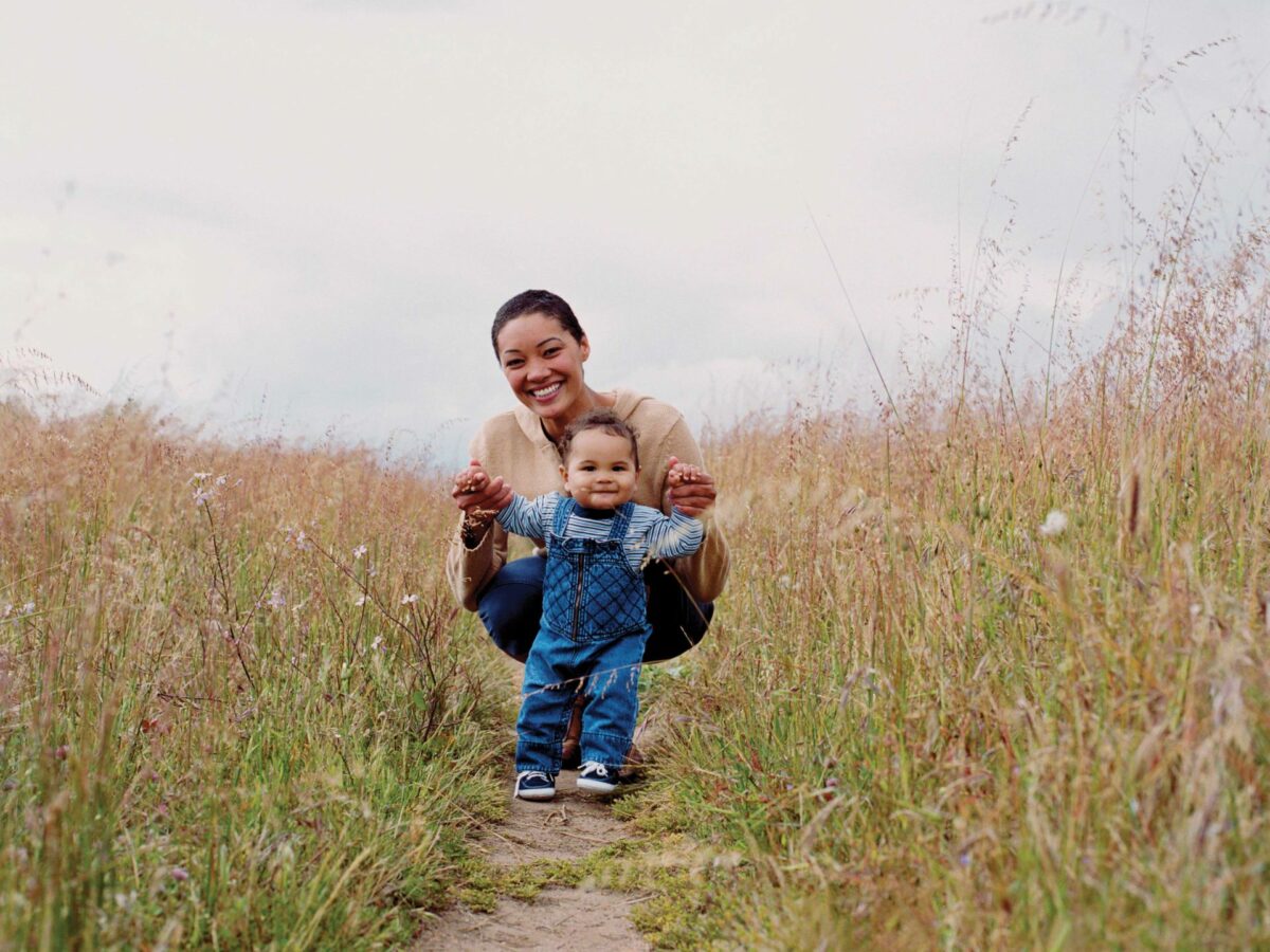 a mother and her child, about 10 months old, pose for a photo on a path with tall grass on either side of them