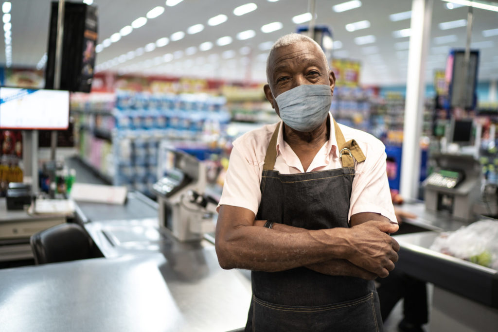 a man wears an employee apron and a mask as he stands with arms folded in a grocery or hardware store