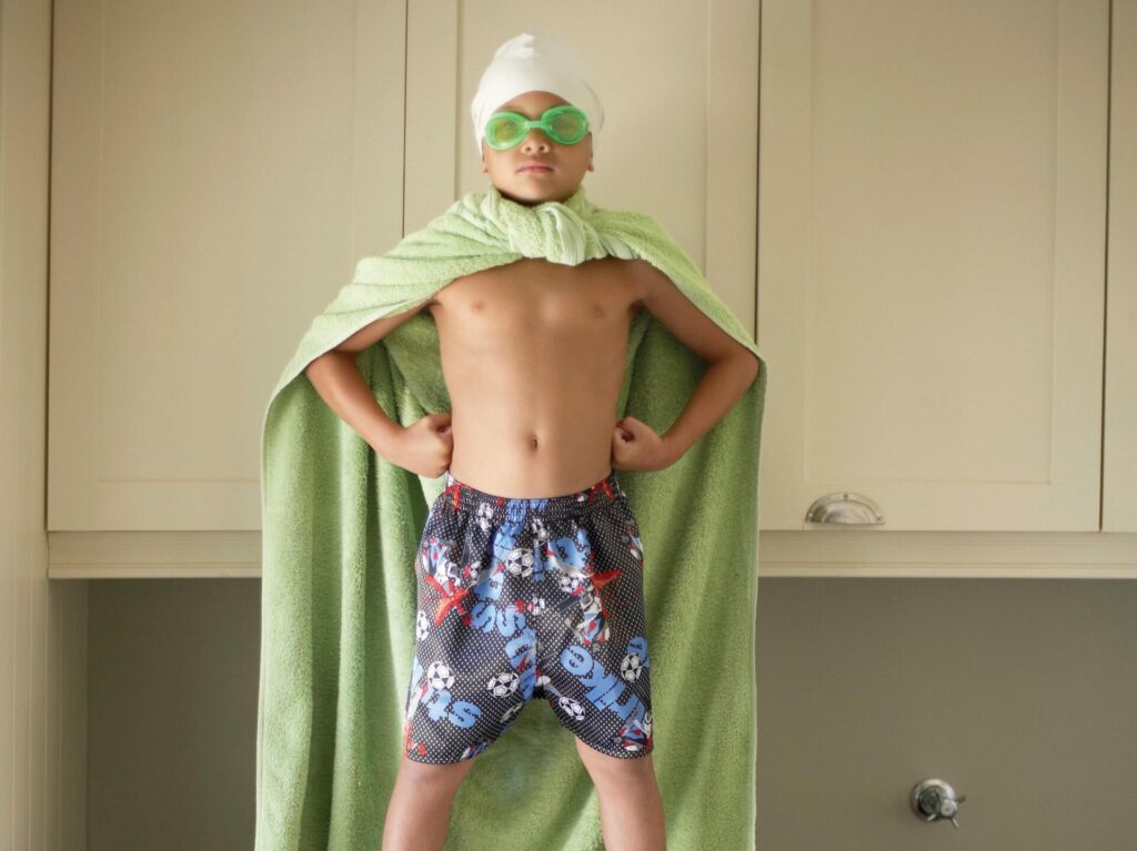A young boy stands on a washing machine and dryer wearing a towel like a cape, swimming goggles and a swimming cap. He is standing with his hands on his hips like a superhero pose.