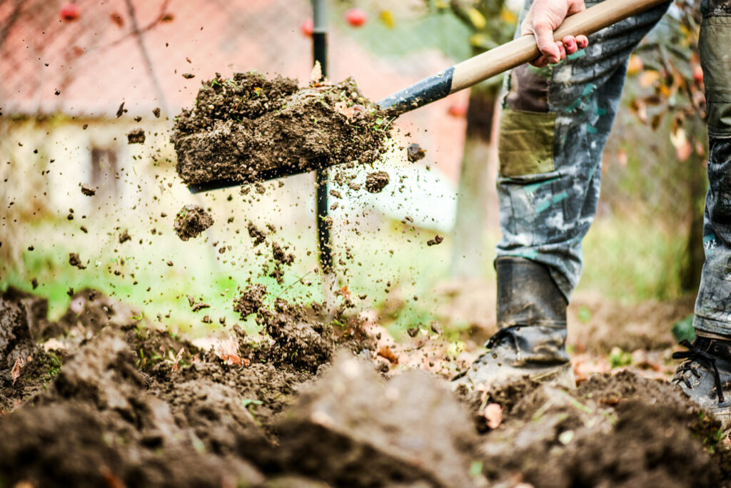 a person lifts a pile of dirt with a shovel. You cannot see the person except their legs and hand on the shovel. The dirt is flying. Call 811 before you dig.