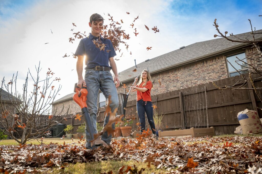 A man uses an electric leaf blower to blow leaves in his back yard. A woman stand behind him tossing leaves with a rake