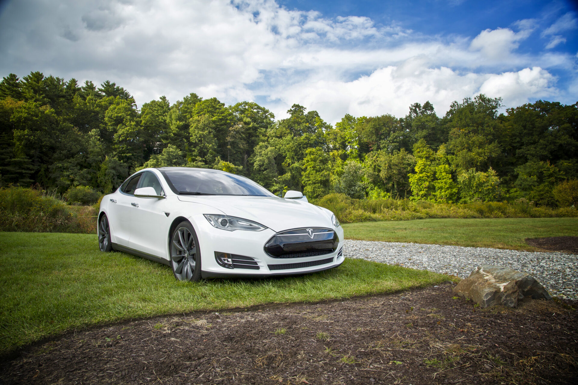 A Tesla electric vehicle is parked on a grassy area next to a gravel road with evergreen trees and blue sky behind it. Photo courtesy of Tesla