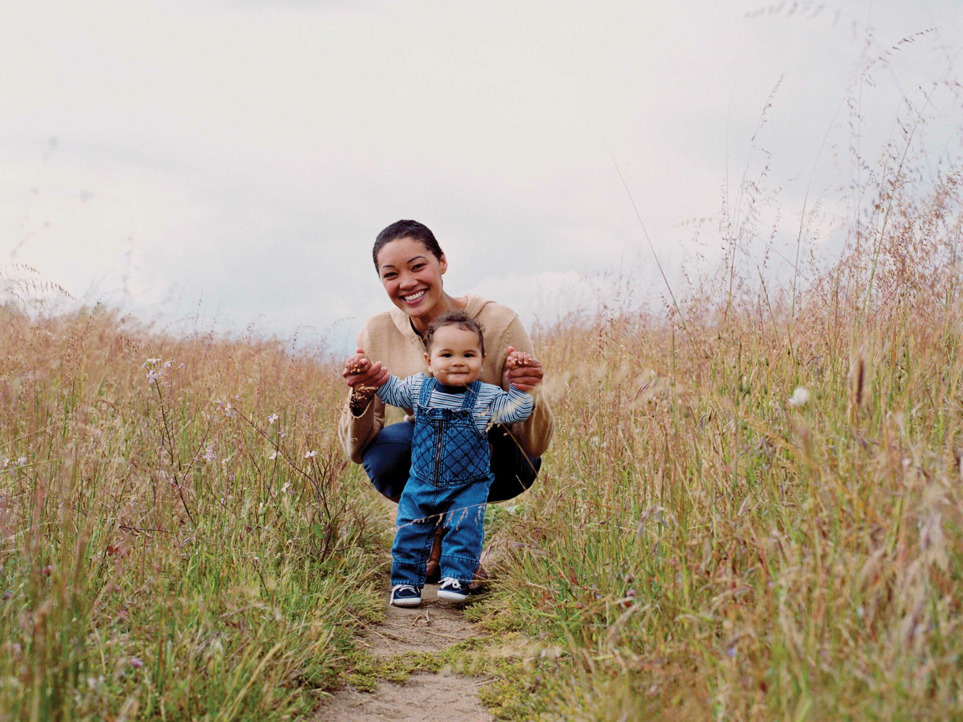 A young mom and her young son crouch together along a trail through tall grass.
