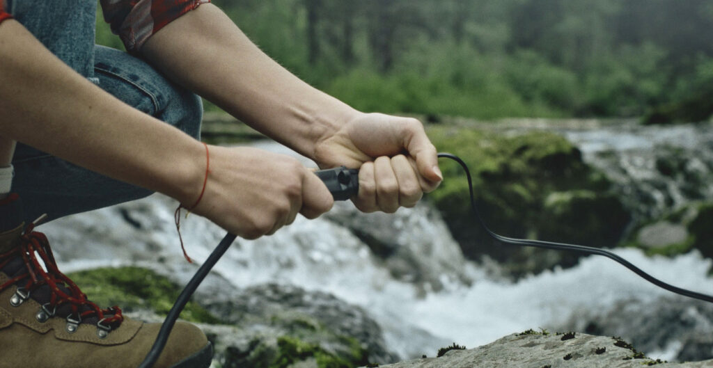 Image of a young Caucasian woman plugging together two power cords with a river flowing behind. The image is zoomed in on her hands. It symbolizes how water and hydropower powers our lives.