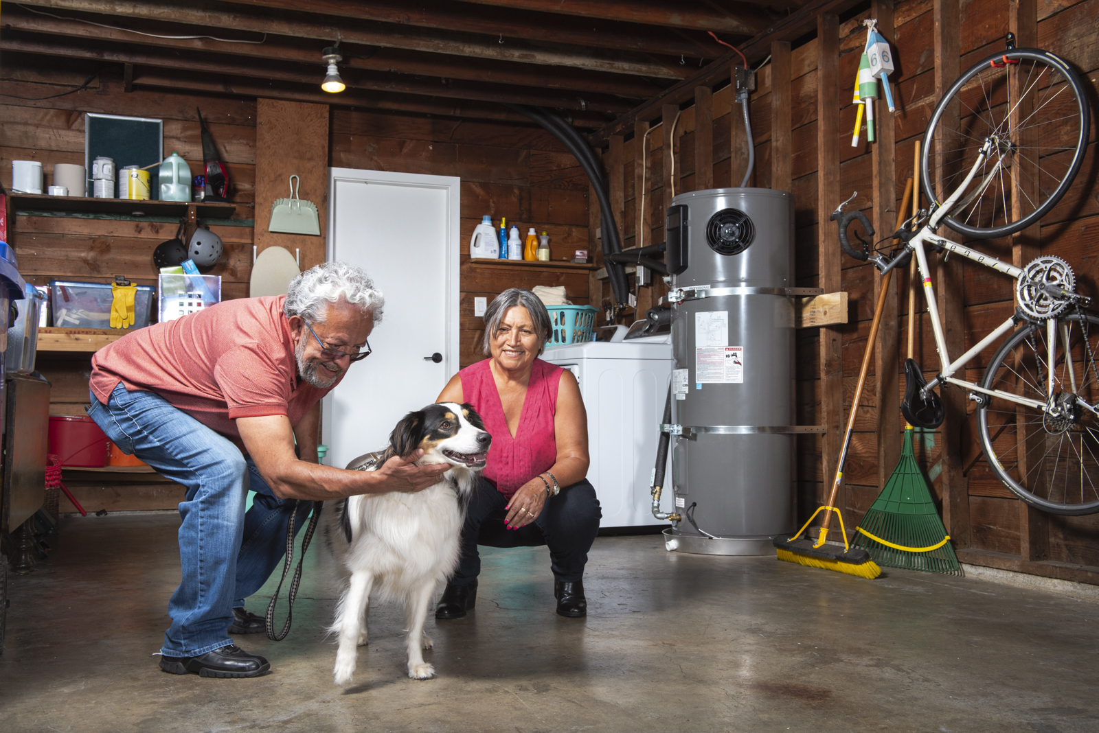 An older couple crouches in the garage to pet their dog. A hybrid electric water heater is in the background saving energy.