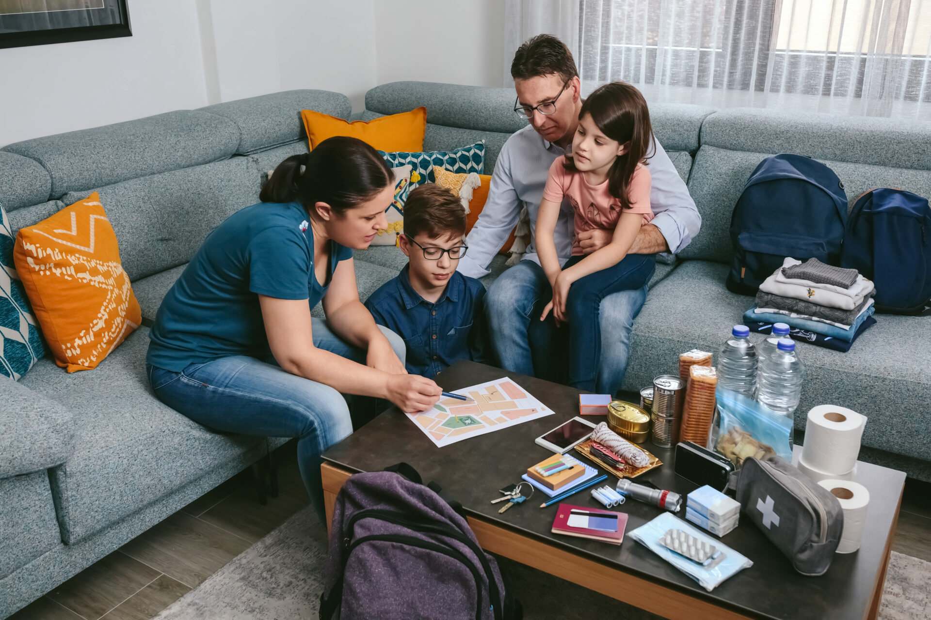 A mother, son, father and daughter sit at a couch looking at a map for their family emergency plan. Emergency supplies and food are on the coffee table. Backpacks and clothes are on the couch and floor.