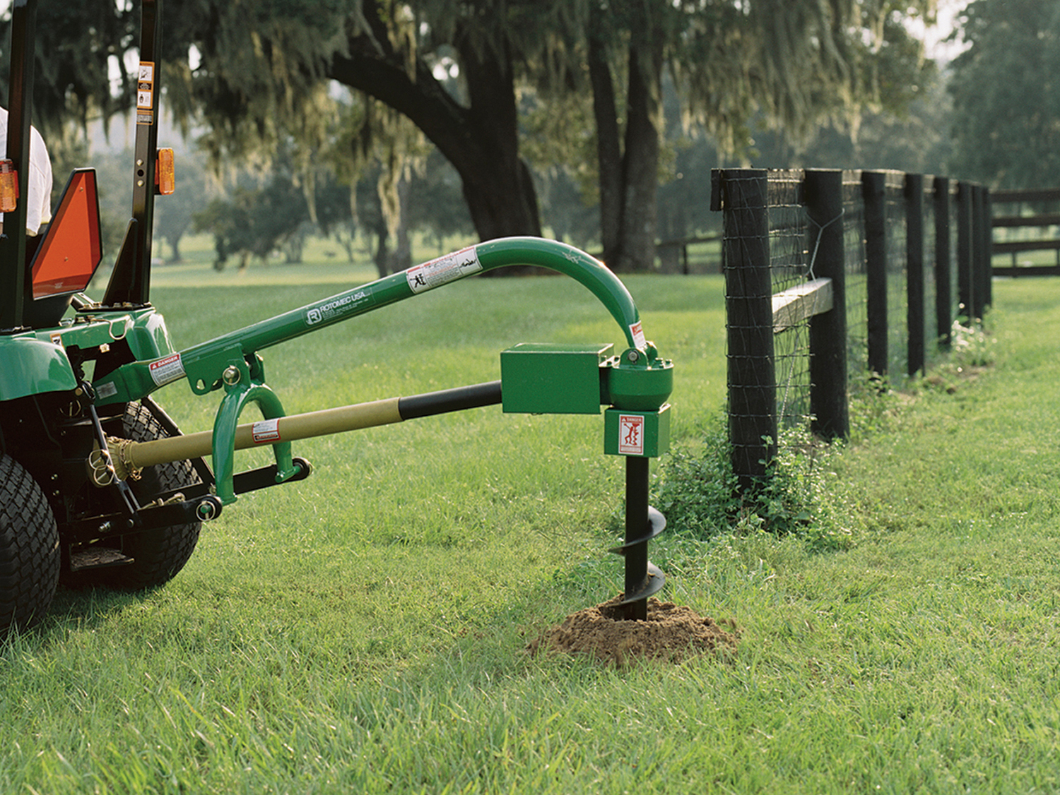 A green lawn tractor digs a post hole along a fence line. Dirt is exposed on a green grassy area. Call 811 before you dig.