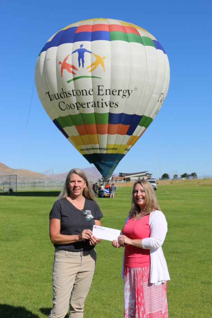 Kristi Thien, nutrition services director with Mid-Columbia Meals on Wheels, accepts a donation from the Cooperative Balloon Associates, presented by Benton REA Board President Connie Krull. Behind them, the Touchstone Energy Cooperatives hot air balloon prepares for a tethered flight. PHOTO BY ELECIA COPENHAVER