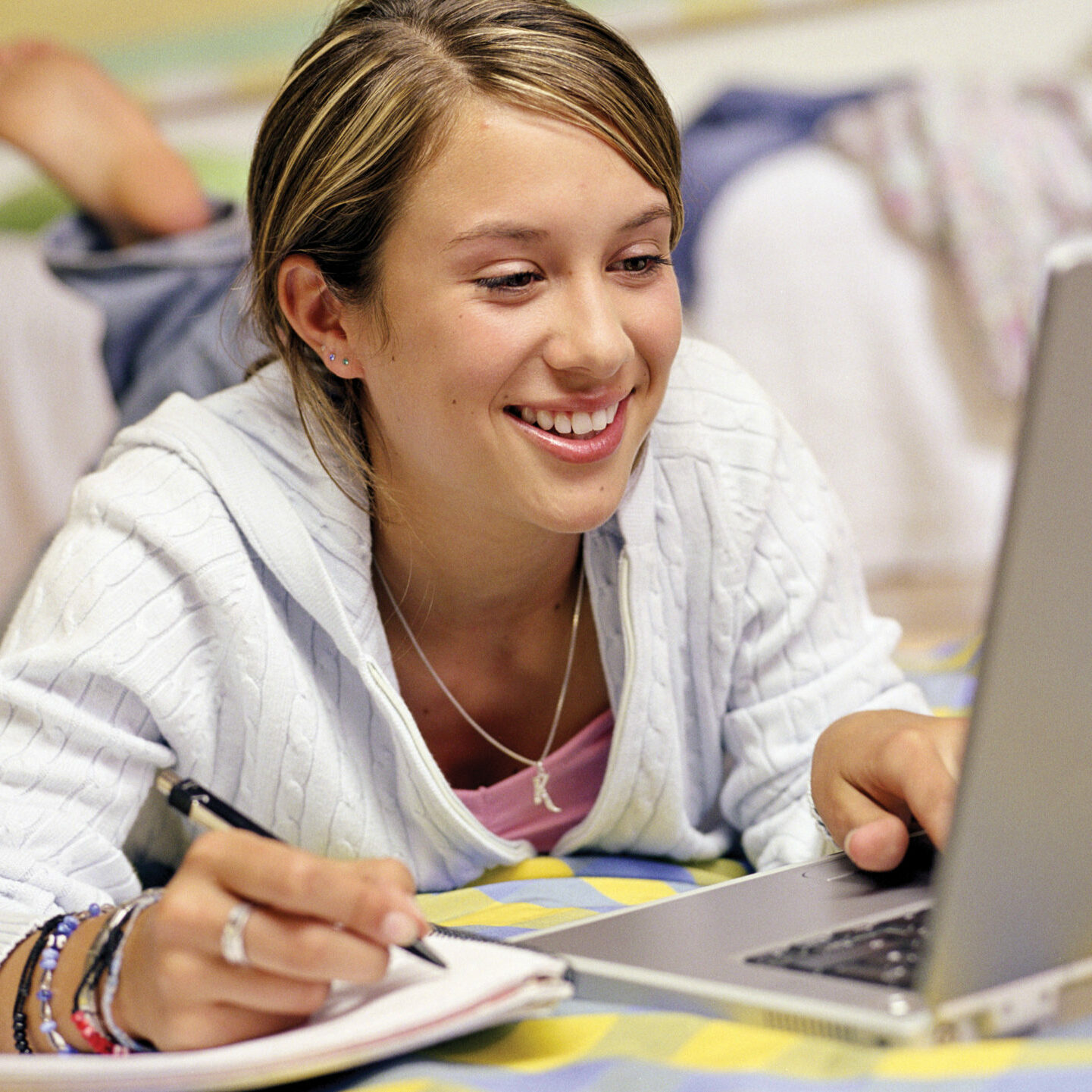 A teenage girl reclines on her bed while looking at a laptop and holding a pen in her right hand about to write in a notebook