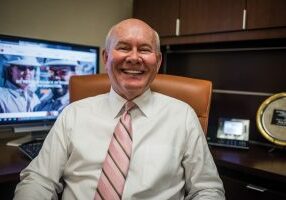Smiling IT Professional wearing a white shirt and pink tie sitting in an office chair at his desk