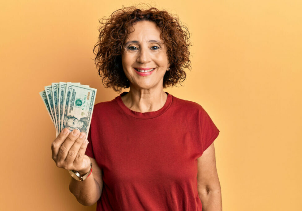 A hispanic woman with curly hair holds several $20 bills in her right hand. She is wearing a red t-shirt and the background is orange.