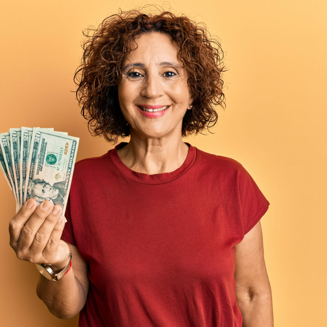 A hispanic woman with curly hair holds several $20 bills in her right hand. She is wearing a red t-shirt and the background is orange.