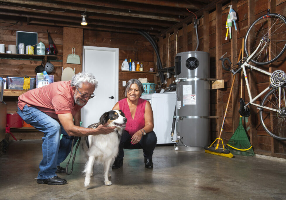 An older couple crouches in the garage to pet their dog. A hybrid electric water heater is in the background saving energy.